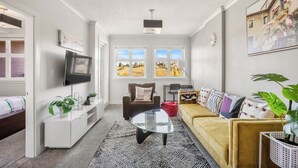 Un salon moderne avec un canapé, un fauteuil et une table basse. Il y a des fenêtres donnant sur des arbres et un tableau accroché au mur.