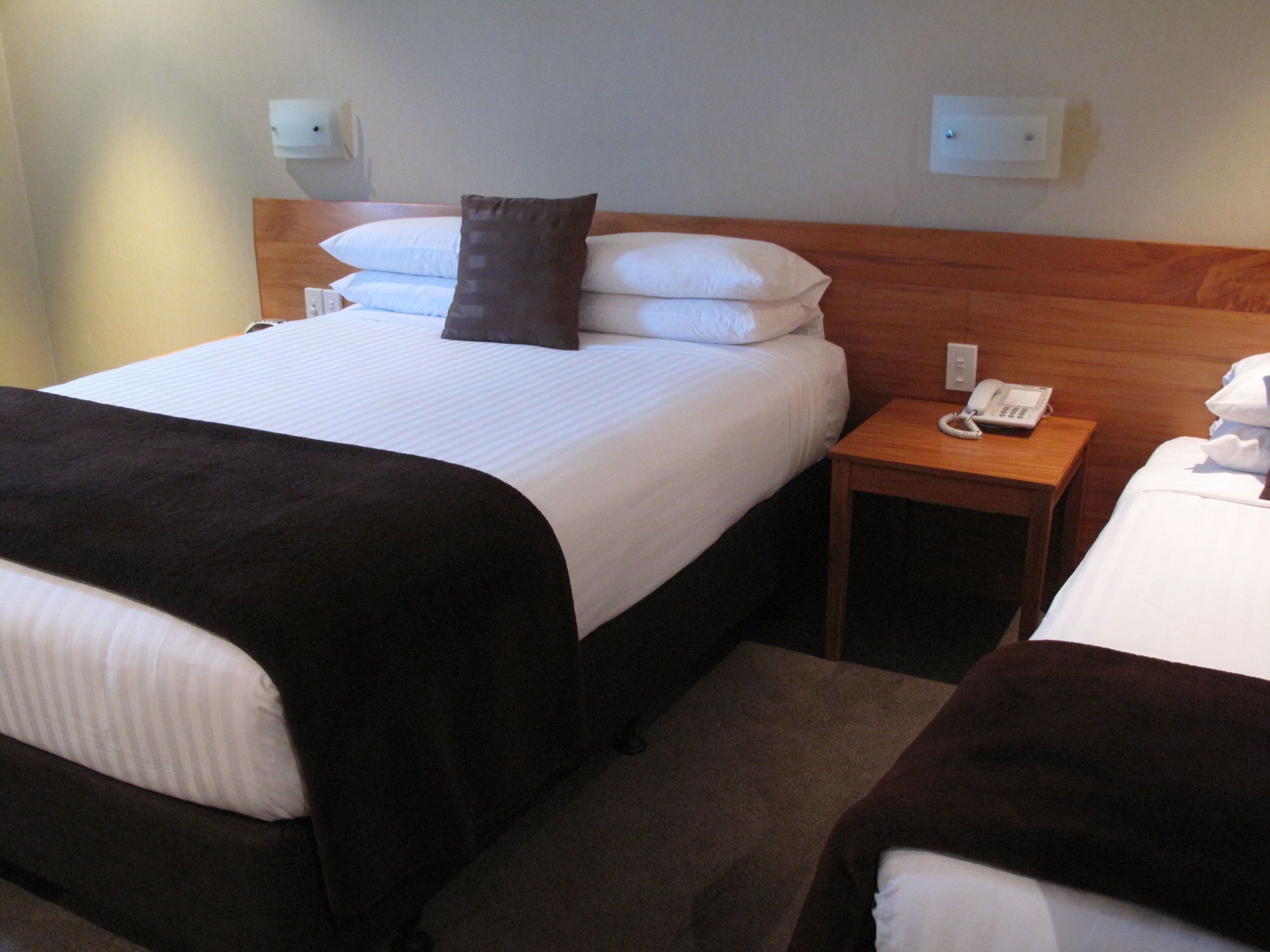 A hotel room with two beds, a wooden headboard, a bedside table with a phone, and a wall socket.