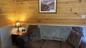 A cozy room with wooden walls, a framed landscape painting, a brown sofa with patterned cushions, and a small table with a lamp and a sign.