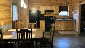 A wooden cabin interior with a dining table, chairs, a refrigerator, a stove, and a window with a view.