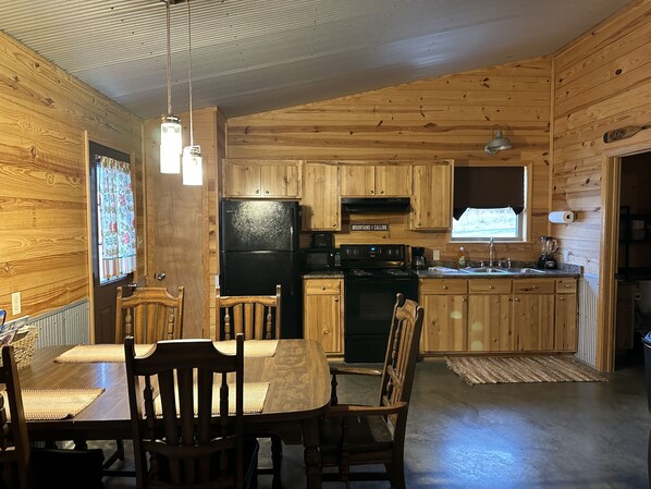 A wooden cabin interior with a dining table, chairs, a refrigerator, a stove, and a window with a view.