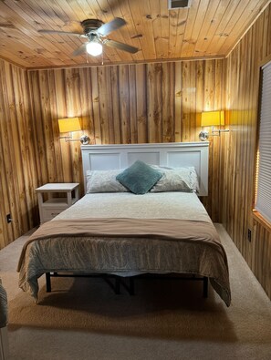 Ein Schlafzimmer mit einer holzgetäfelten Wand, einem Bett mit gemusterten Betttextilien, einem Deckenventilator und zwei Wandlampen.