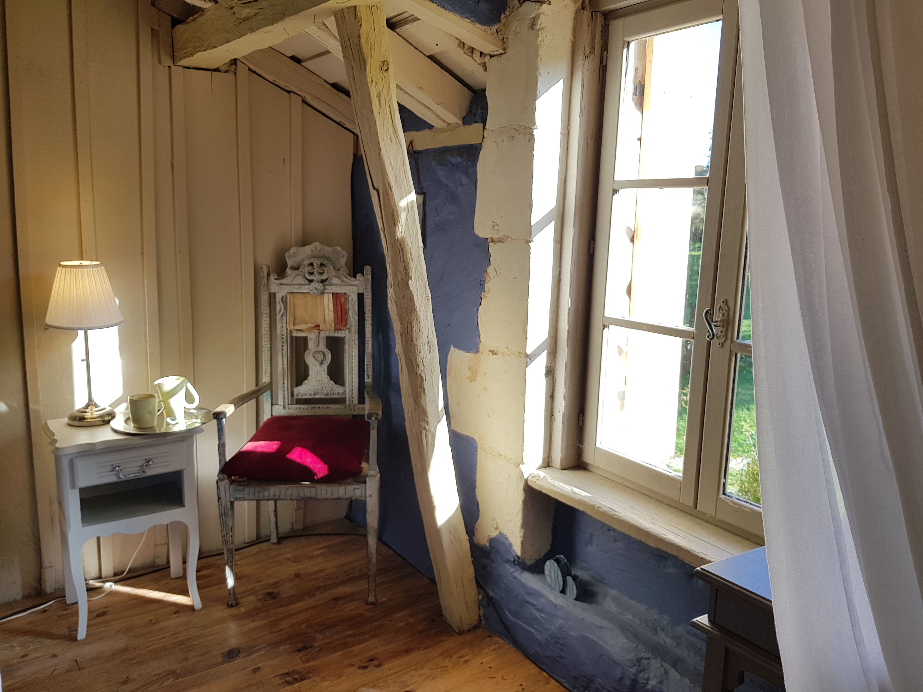 Una habitación con piso de madera, una silla blanca con cojín rojo, una mesita con lámpara y una ventana con cortinas.
