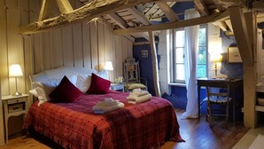 A rustic bedroom with a wooden ceiling, a bed with red bedding, a nightstand with a lamp, and a desk with a lamp.