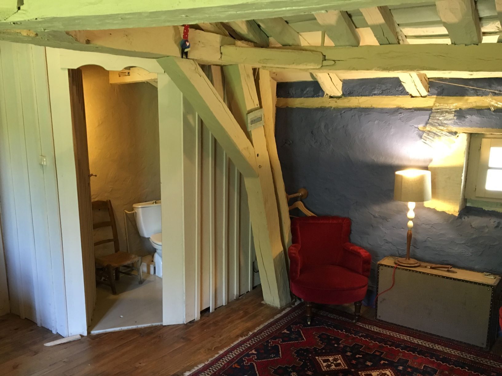 A rustic room with a wooden staircase, a red chair, a small table with a lamp, and a patterned rug.