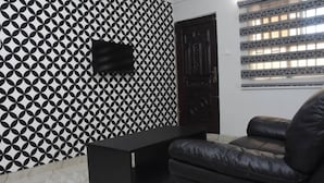 A room with a geometric patterned wall, a black TV mounted on the wall, a dark wooden door, and a black leather sofa. - Beautiful 1 Bed, Living Room in a Hotel Penthouse (Awoyaya, Ibeju Lekki LG.)