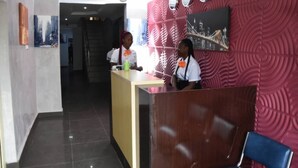A reception area with a wooden reception desk, a red textured wall, and a cityscape painting. - Beautiful 1 Bed, Living Room in a Hotel Penthouse (Awoyaya, Ibeju Lekki LG.)