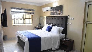 A bedroom with a large bed, a nightstand, a wall-mounted air conditioner, a window with bars, and a cityscape painting. - Beautiful 1 Bed, Living Room in a Hotel Penthouse (Awoyaya, Ibeju Lekki LG.)