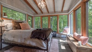 Een slaapkamer met een groot bed, een houten bankje en uitzicht op bomen door grote ramen.