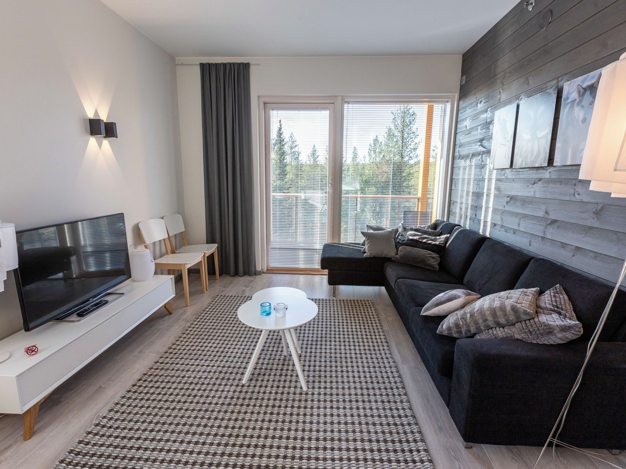 A modern living room with a flat-screen TV, a sofa, a coffee table, and a large window with blinds.