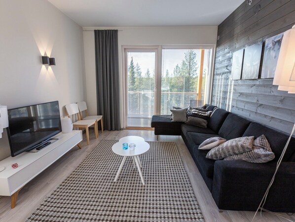 A modern living room with a flat-screen TV, a sofa, a coffee table, and a large window with blinds.