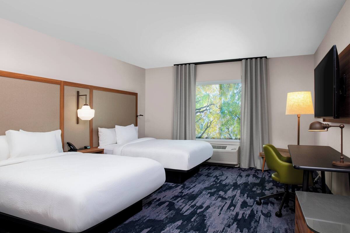 Ein Hotelzimmer mit zwei Betten, einem Schreibtisch, einem Stuhl, einem Fernseher, einer Lampe und einem Fenster mit Vorhängen.