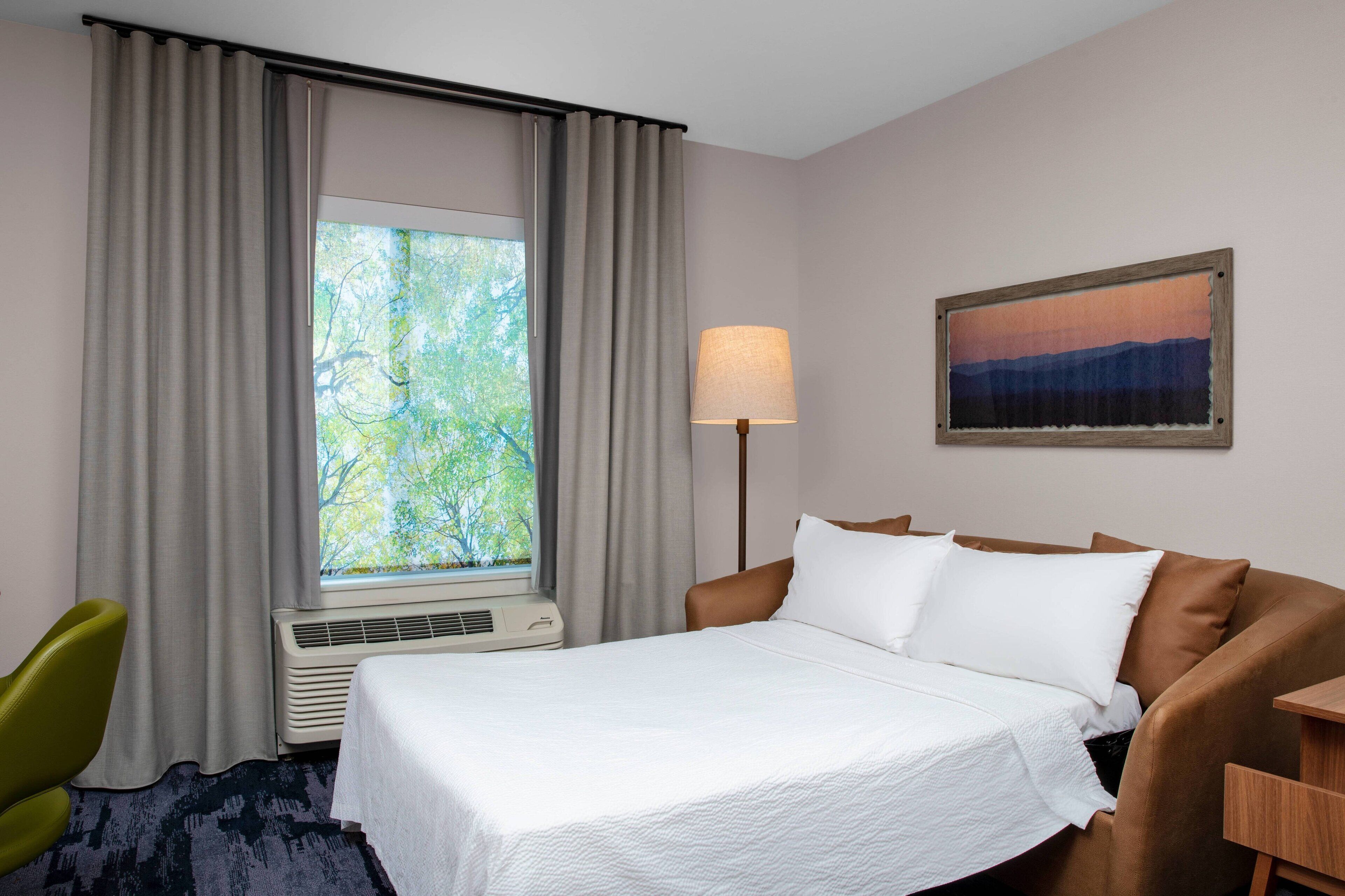 A hotel room with a bed, a desk, a chair, a window with curtains, a wall-mounted lamp, and a painting.