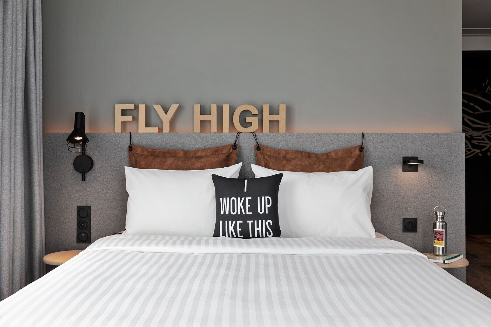 A neatly made bed with striped bedding, a pillow with motivational text, a wall-mounted light fixture, and a decorative wall piece.