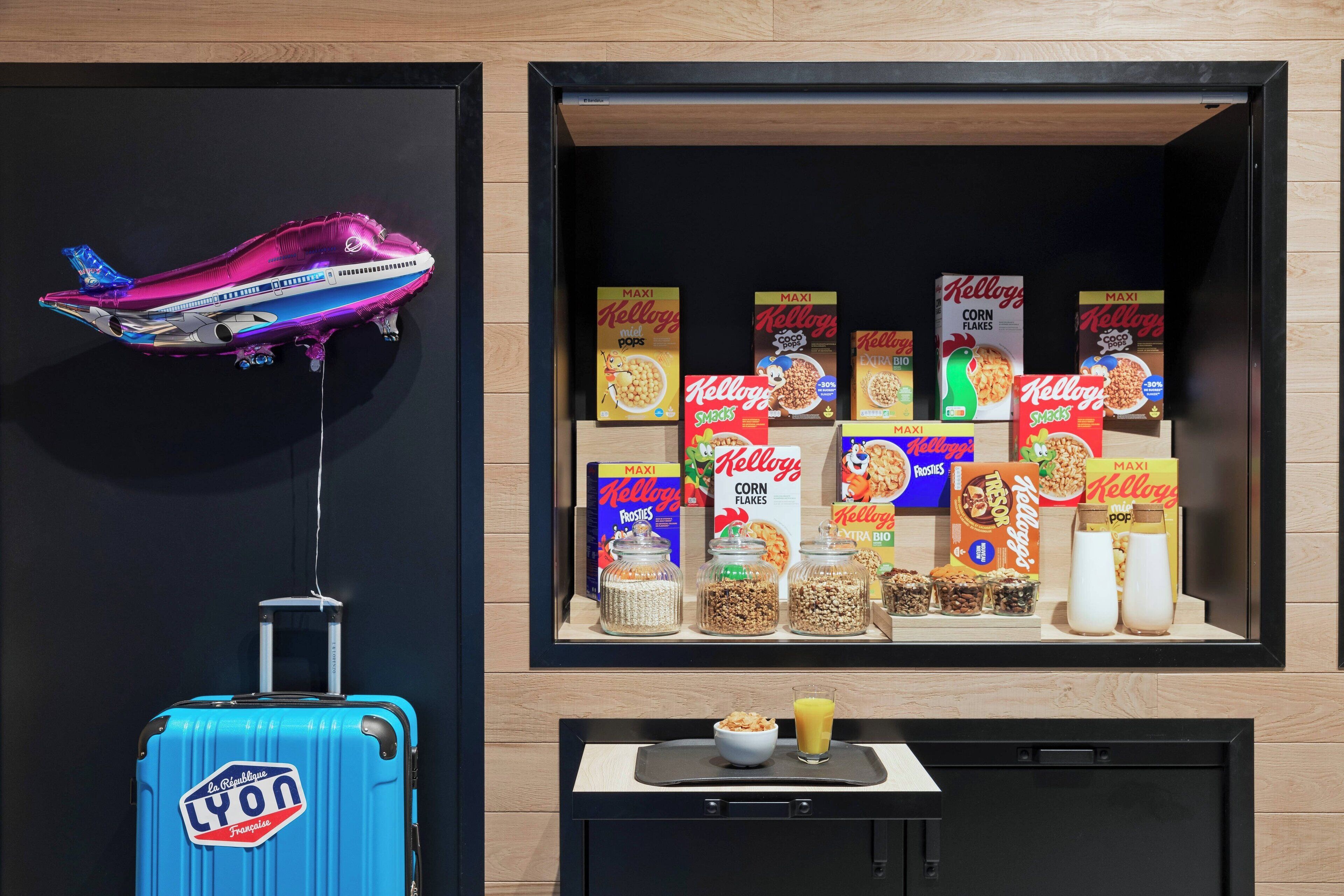 A room with a luggage trolley, a blue suitcase, and a wall display of Kellogg's cereals.