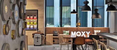 A modern interior with a dining area, a sofa labeled "MOXY," and a wall displaying various items.