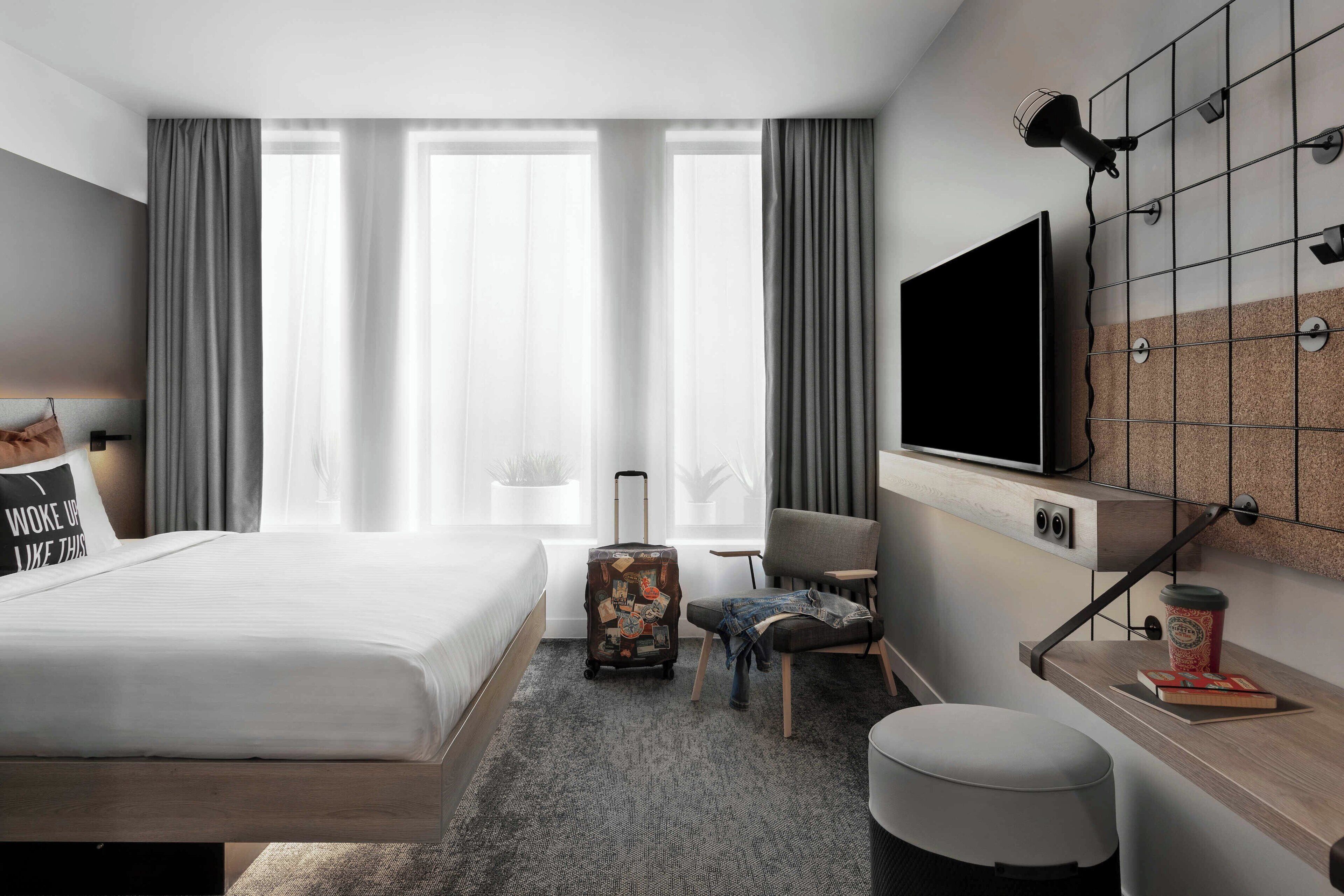 Una habitación de hotel moderna con una cama grande, un escritorio con televisor de pantalla plana, una silla, una maleta y una taza de café.