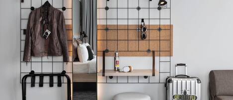 Una habitación de hotel moderna con un panel de pared enrejado, un perchero colgante, un espejo, un estante pequeño con una cámara y una botella, una maleta con una etiqueta que dice 'Eats Sleep Fly', y un taburete redondo.
