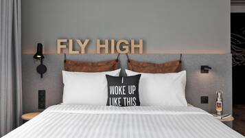 En pænt redt seng med stribet sengetøj, en pude med motiverende tekst, en vægmonteret lampe og et skilt med teksten "FLY HIGH".