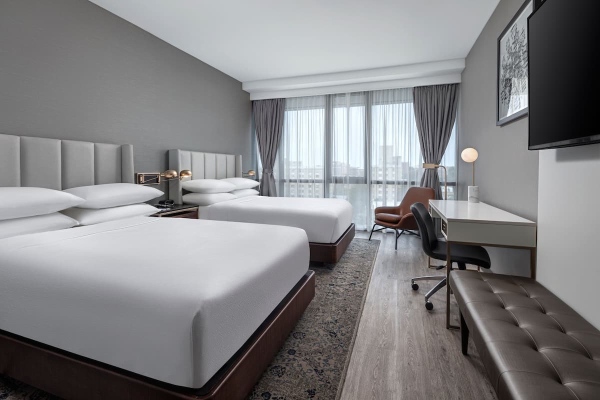 Una habitación de hotel moderna con dos camas, un escritorio, una silla, un banco y un amplio ventanal con cortinas.
