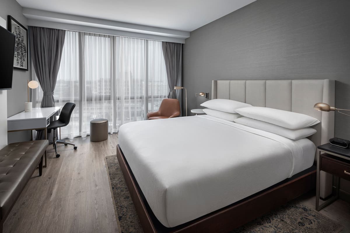 Habitación de hotel moderna con una cama grande, un escritorio, una silla y vistas de la ciudad a través de cortinas translúcidas.