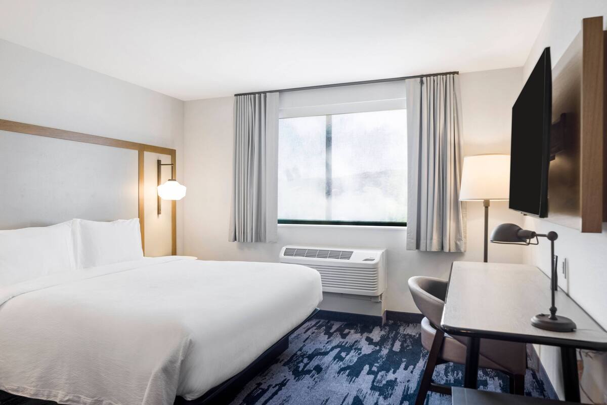 A hotel room with a large bed, a desk, a chair, a TV, and a window with curtains.