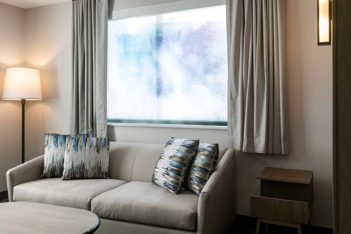 A modern hotel room with a sofa, a lamp, a small wooden side table, and a window with curtains.