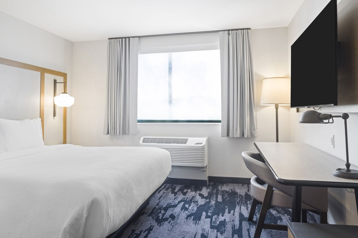 Habitación de hotel con cama, escritorio, silla, televisor, lámpara, aire acondicionado y ventana con cortinas.
