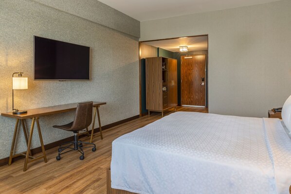 Habitación de hotel con cama, escritorio con silla, televisor montado en la pared y armario.