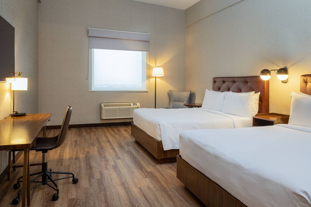 Hotelkamer met twee bedden, een bureau en een stoel. Er is een raam met jaloezieën, een staande lamp en een wandradiator.