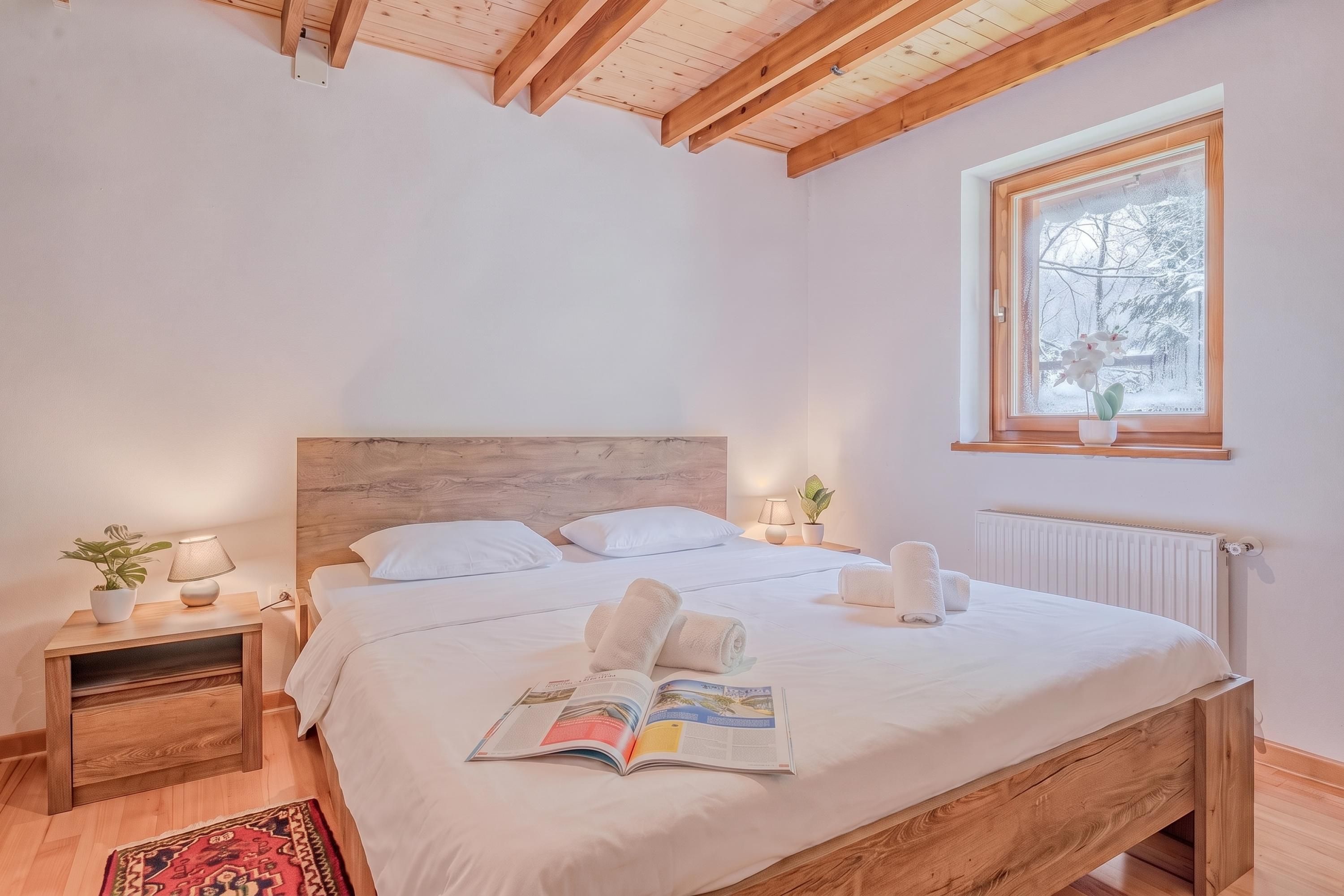 Ein Schlafzimmer mit einem Holzbett, weißer Bettwäsche, einem Fenster mit Aussicht, einem Nachttisch mit Lampe und einer Zeitschrift auf dem Bett.
