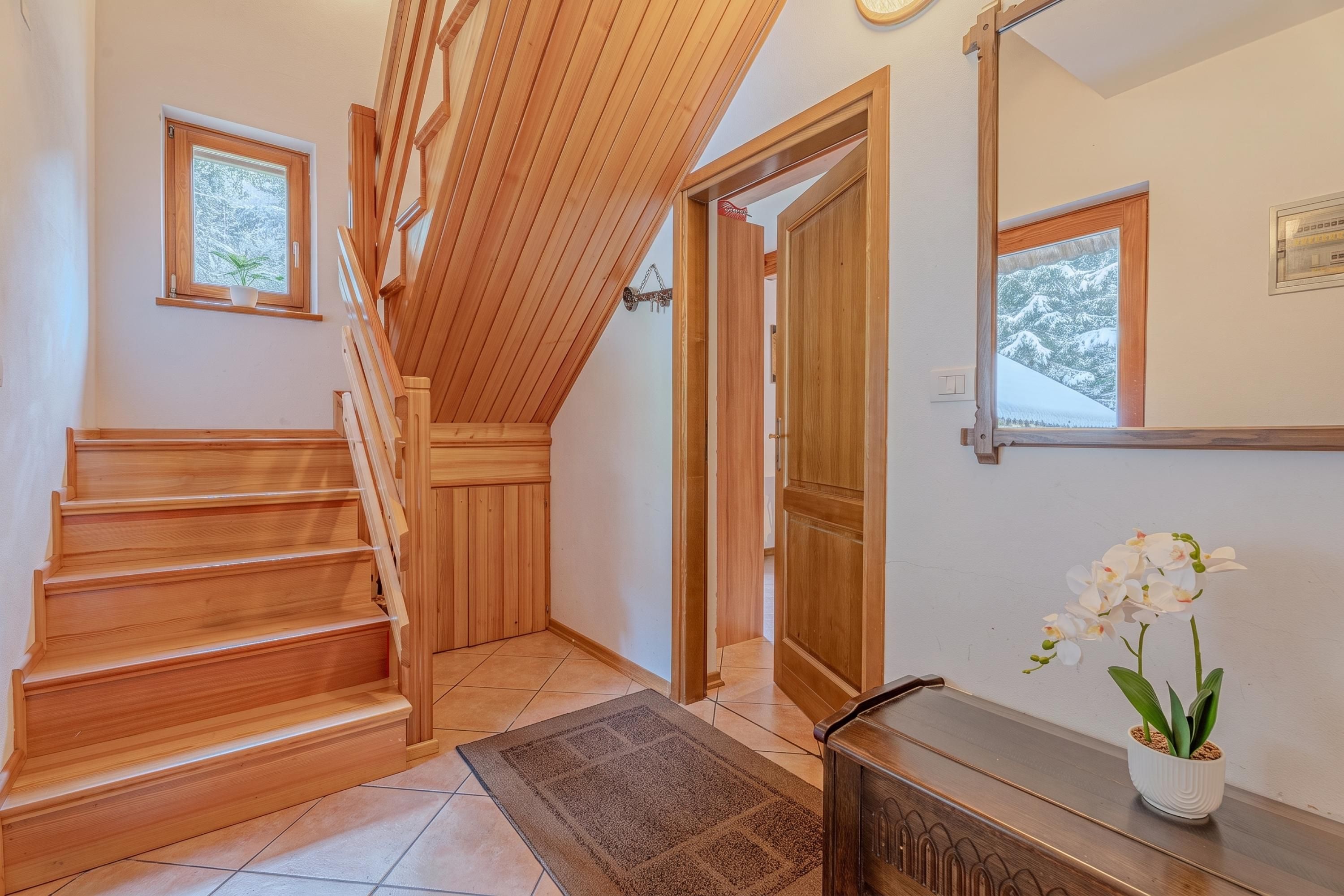 A wooden staircase leading to a second floor, a door with a view of snow-covered trees, and a potted orchid on a console table.