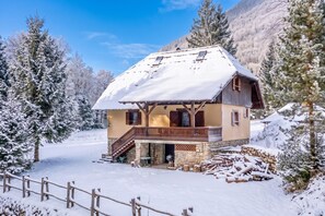 A chalet with a balcony, stone foundation, and a snow-covered roof, surrounded by snow-laden trees.