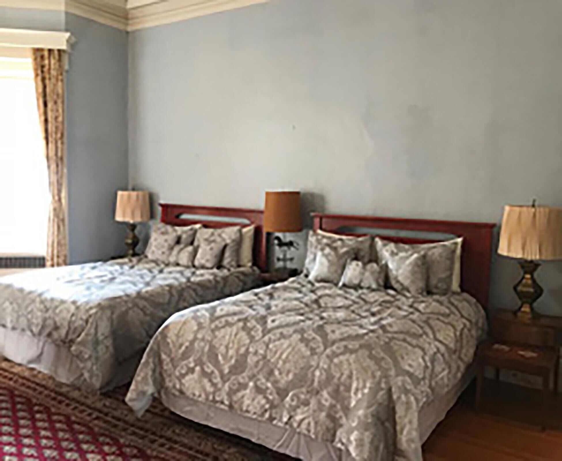 A bedroom with two beds, each with a headboard, a nightstand with a lamp, and a window with curtains.