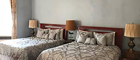 A bedroom with two beds, each with a headboard, a nightstand with a lamp, and a window with curtains.