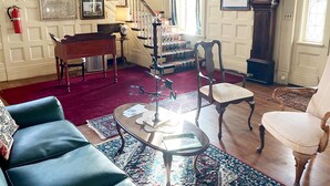 Een ruime woonkamer met een statige trap, een piano, een kroonluchter en een grote klok.