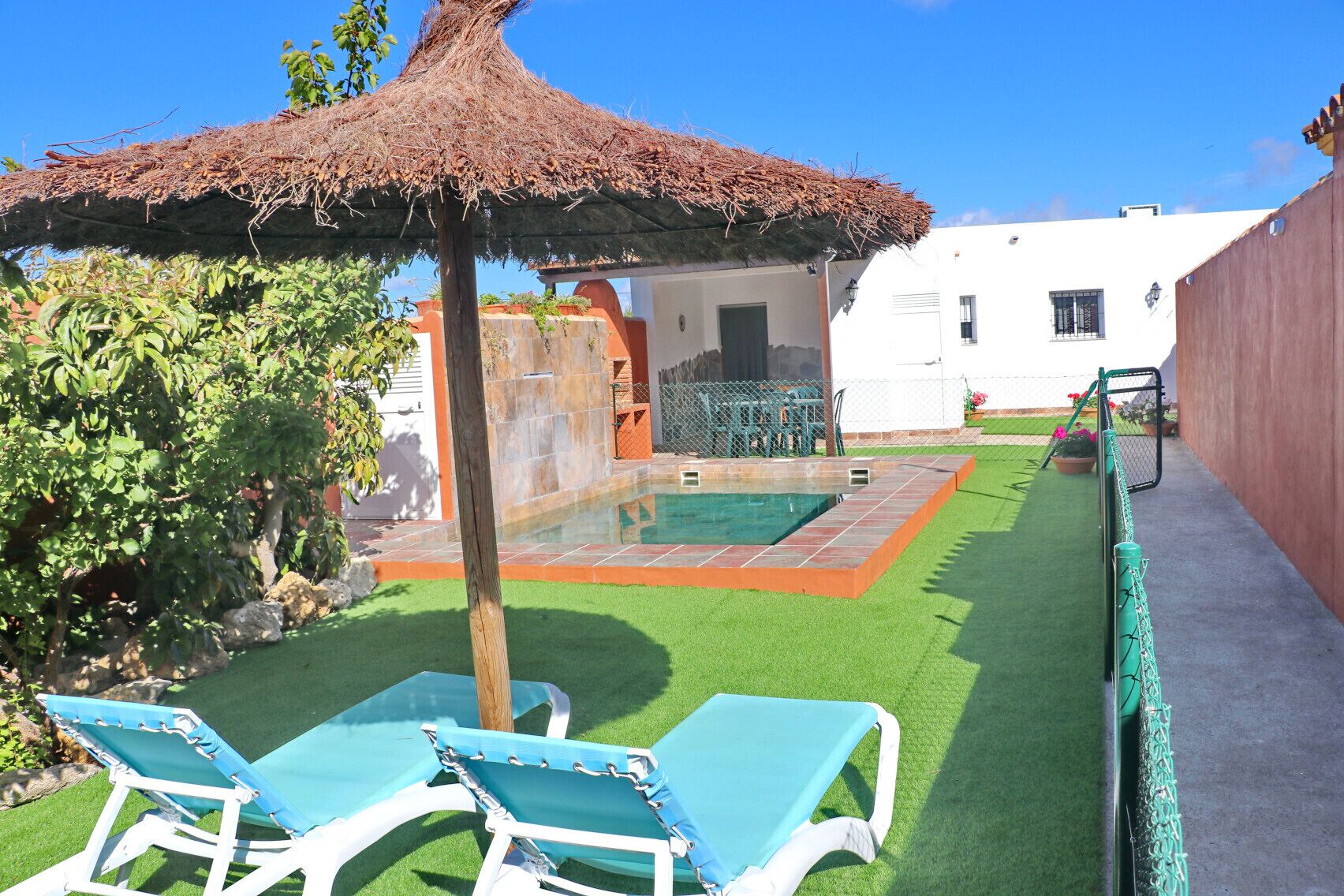 Un jardin avec une piscine, deux chaises longues, un parasol en chaume et un espace clos agrémenté de plantes en pot.