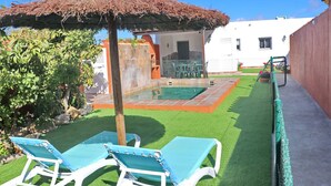Un jardin avec une piscine, deux chaises longues, un parasol en chaume et un espace clos agrémenté de plantes en pot.