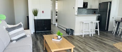 A modern living room with a wooden coffee table, a grey sofa, and a black wicker console. A kitchen with white cabinets and a black refrigerator is visible in the background.