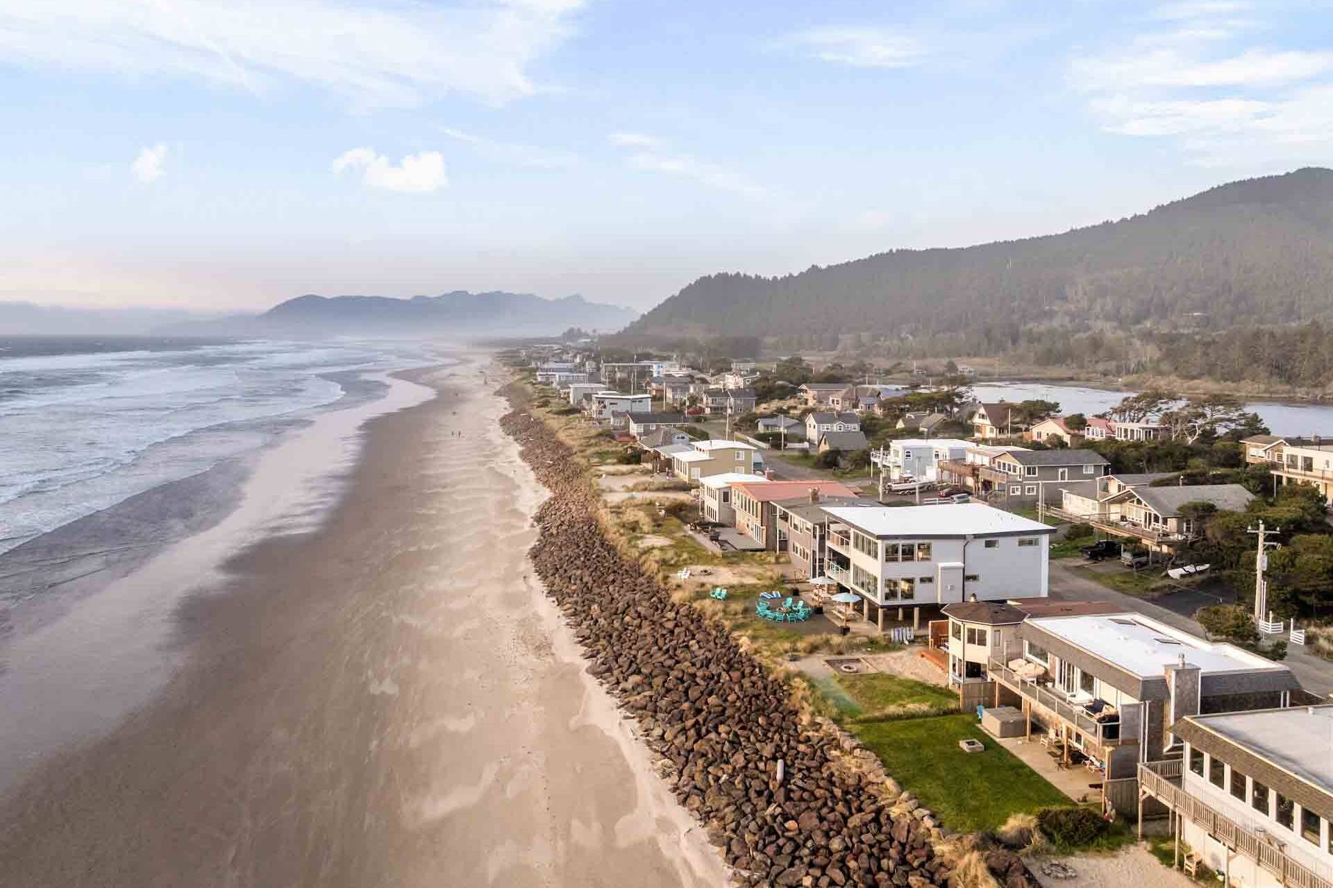 A coastal residential area with houses along the beach, a rocky shoreline, and a hilly backdrop.