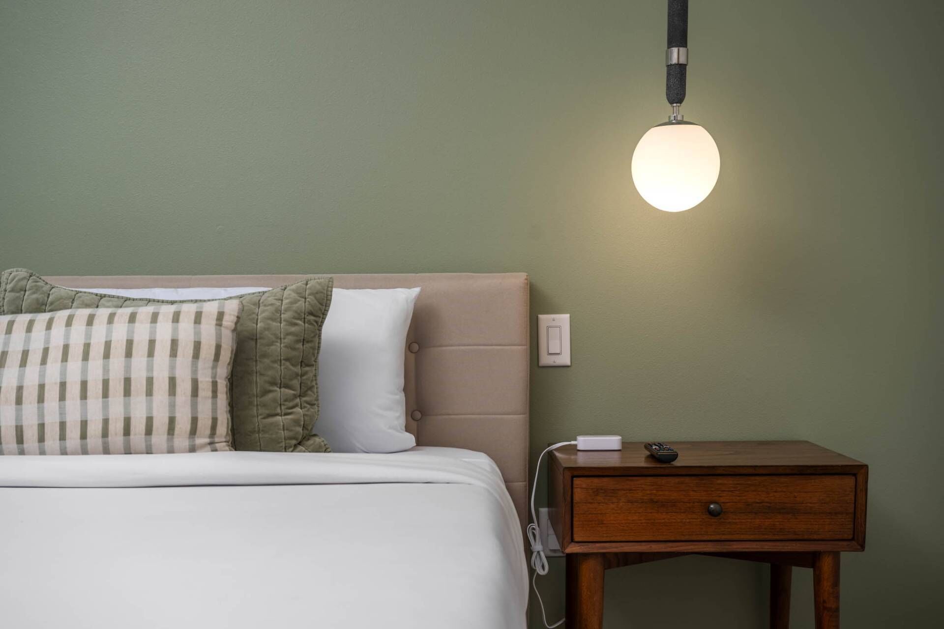 Un letto rifatto con un cuscino a quadretti, un comodino con telecomando e ciabatta multipresa, e un lampadario fissato al muro.