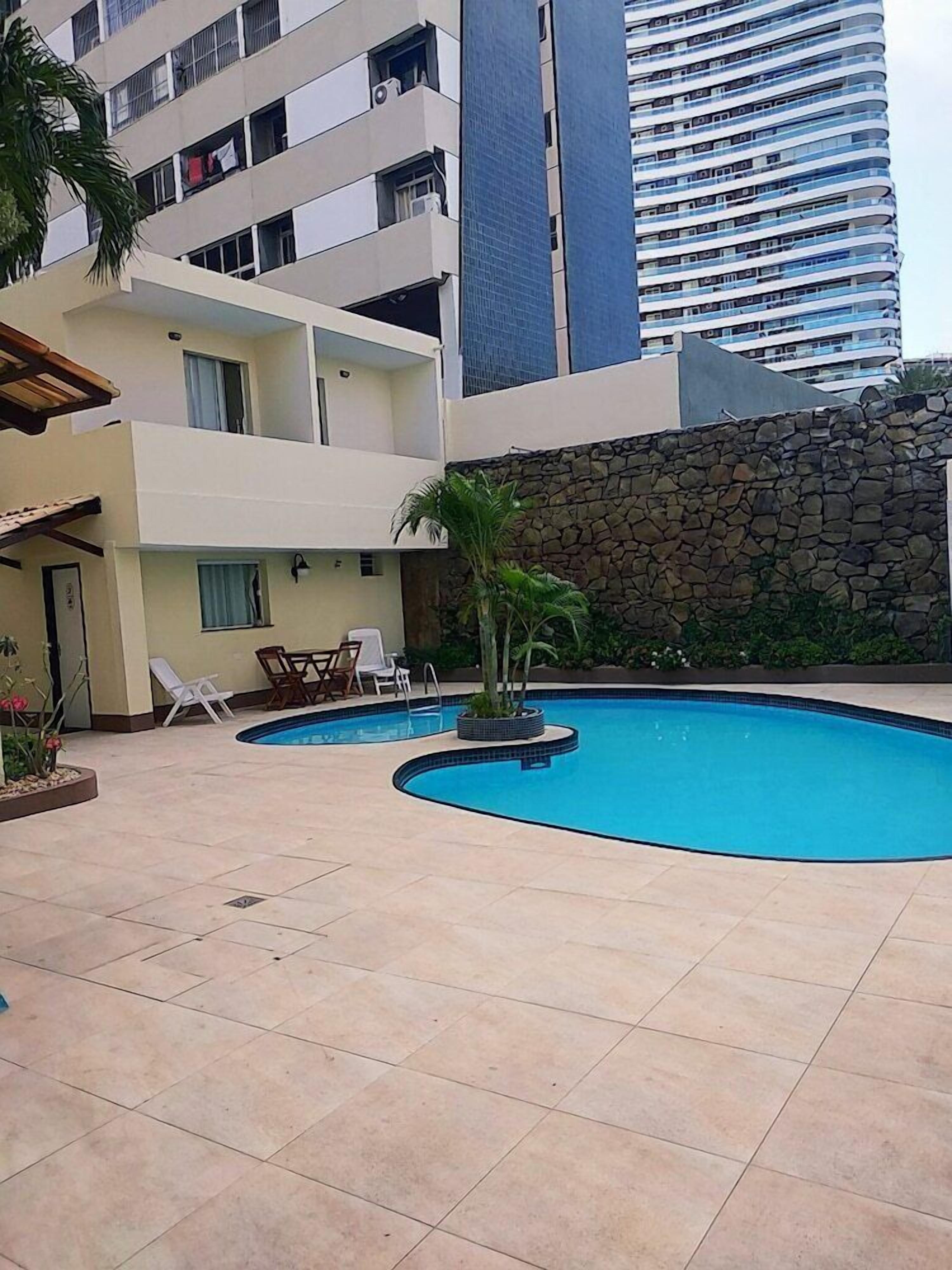 Uma piscina com espreguiçadeiras e uma parede de pedra, cercada por um edifício com varandas e uma estrutura alta ao fundo.