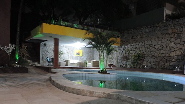 Un espace piscine nocturne avec un mur en pierre, une structure jaune et un coin salon comprenant une chaise et une table.