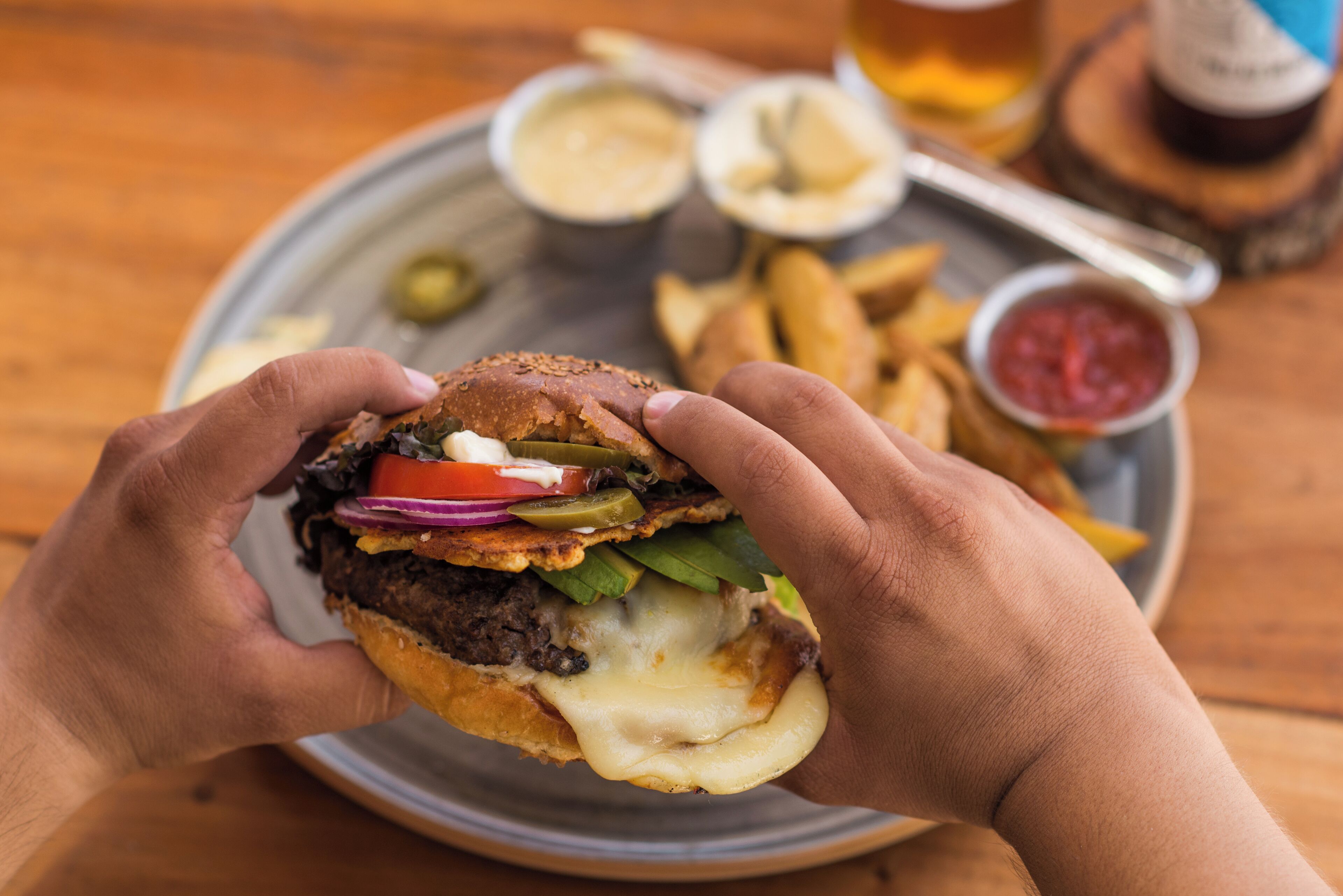 A person holding a burger with various toppings and a plate of fries and dipping sauces.