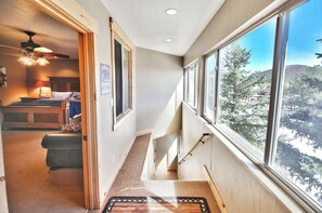Ein schmaler Flur mit Teppichboden, einem Bett mit Kopfteil, einem Deckenventilator und einem Fenster mit Blick auf Bäume und einen Berg.