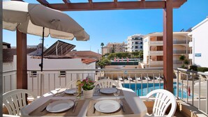 An outdoor dining area with a table set for two, a pool in the background, and a view of a residential building. - Apartamento Água Brisa with Pool, Wi-Fi, A/C, Balcony & Garden (Albufeira)