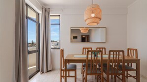 A dining area with a table and chairs, a hanging light fixture, a mirror on the wall, and a view through a window.