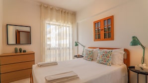 A neatly made bed with a white duvet, a wooden dresser, a small table with a lamp, and a window with curtains.
