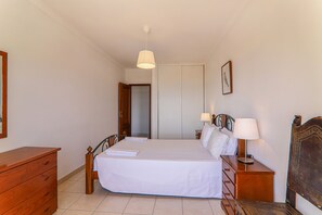 A bedroom with a bed, bedside tables, a wooden dresser, a hanging lamp, and a door leading to another room.