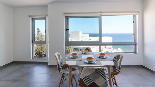 Ein Essbereich mit einem Tisch für vier Personen, großen Fenstern mit Meerblick und einem Balkon mit Pflanze.